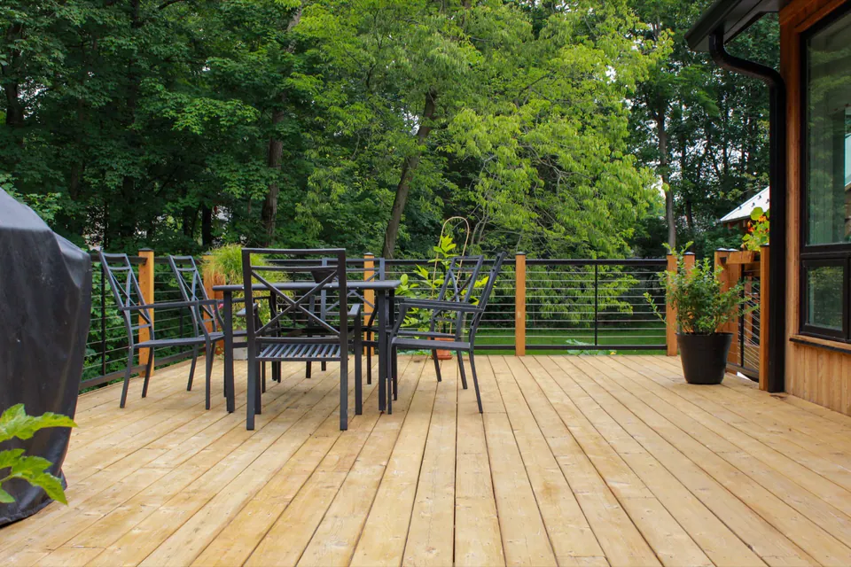 Wood deck dining area with black cable railing, grill, patio furniture, and wooded view.